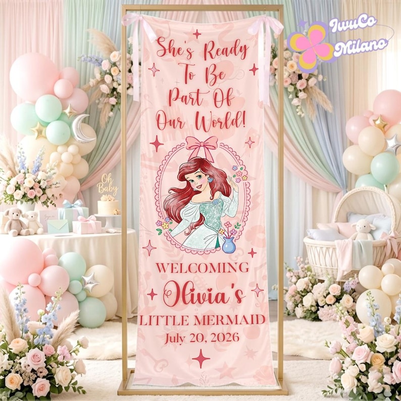May include: A pink banner with the text "She's Ready To Be Part Of Our World!" and "Welcoming Olivia's Little Mermaid July 20, 2026." It features an illustration of Ariel from The Little Mermaid. The banner is part of a baby shower setup.