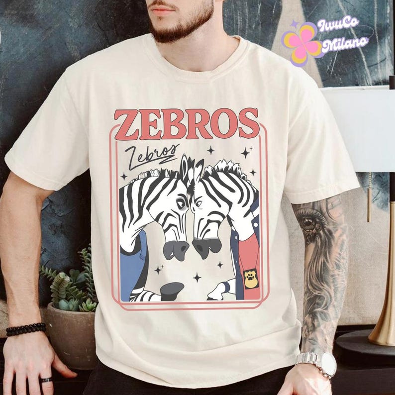 May include: Cream-colored t-shirt featuring a graphic of two zebras with black and white stripes, wearing blue and red jackets. The text "ZEBROS" is above the zebras in a red font, with "Zebros" in cursive below. The shirt is a casual, unisex style.