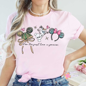 May include: Pale pink t-shirt with a cartoon design featuring Mickey Mouse ears, Olaf the snowman, and a pink bow. The text "For the first time in forever" is printed below the design. A fun, casual top.