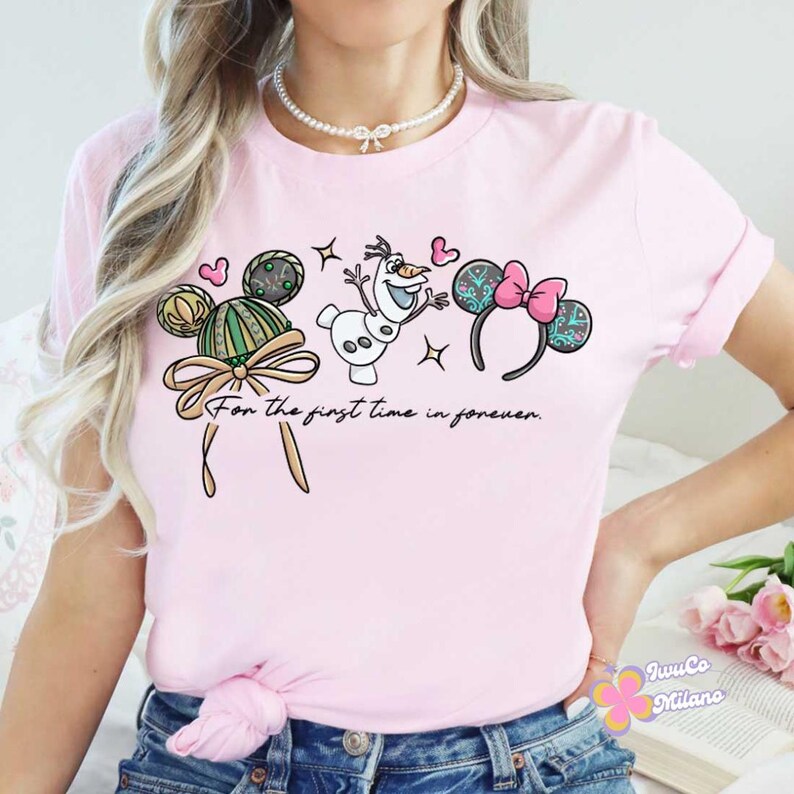 May include: Pale pink t-shirt with a cartoon design featuring Mickey Mouse ears, Olaf the snowman, and a pink bow. The text "For the first time in forever" is printed below the design. A fun, casual top.