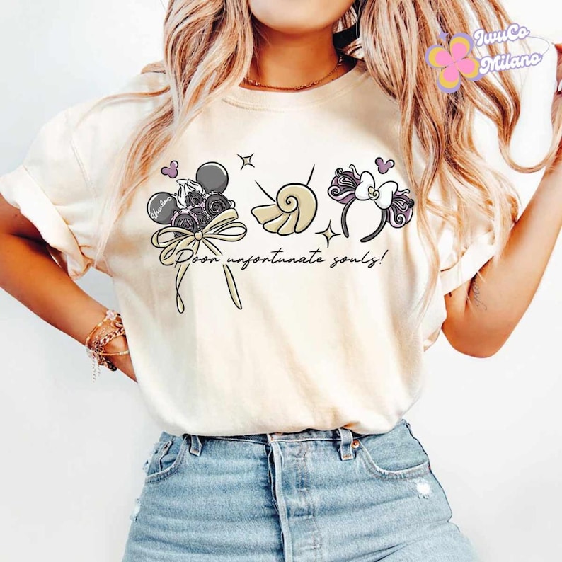 May include: Cream-colored t-shirt featuring a whimsical design with the text "Poor unfortunate souls!". The graphic includes a bow, a seashell, and a headband with mouse ears. The design incorporates black, purple, and beige elements.