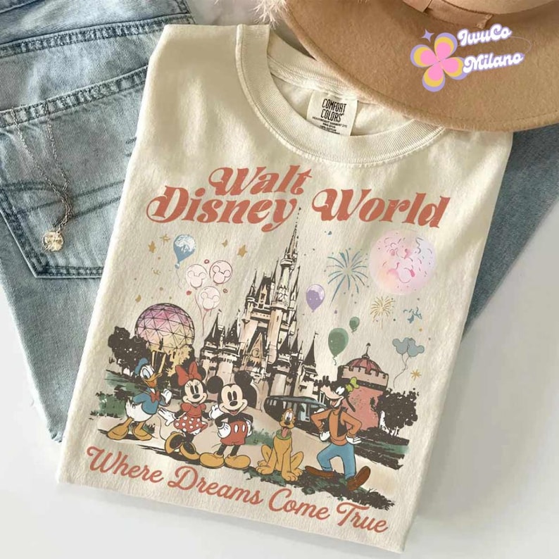 May include: Cream-colored Walt Disney World t-shirt featuring a graphic of the Disney castle, balloons, fireworks, and Disney characters. The text "Walt Disney World" and "Where Dreams Come True" are printed on the shirt. A brown hat and blue jeans are in the background.