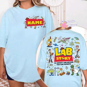 May include: Light blue t-shirt with a red rectangle that says "NAME" in yellow. The back of the shirt has the words "LAB STORY" and "TO INFINITY AND BEYOND FOR OUR PATIENTS!" with cartoon characters and science-themed graphics.