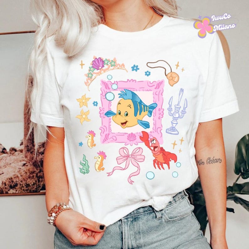 May include: White t-shirt featuring a colorful cartoon design with a yellow and blue fish in a pink frame, a red crab, and other sea-themed illustrations. The shirt has the text "IwuCo Milano" in a floral design.