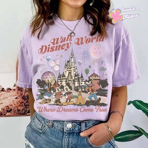 May include: A lilac-colored t-shirt featuring a vintage-style graphic of Cinderella's castle, Mickey and Minnie Mouse, Donald Duck, Goofy, and Pluto. The text "Walt Disney World" and "Where Dreams Come True" are printed above and below the characters.