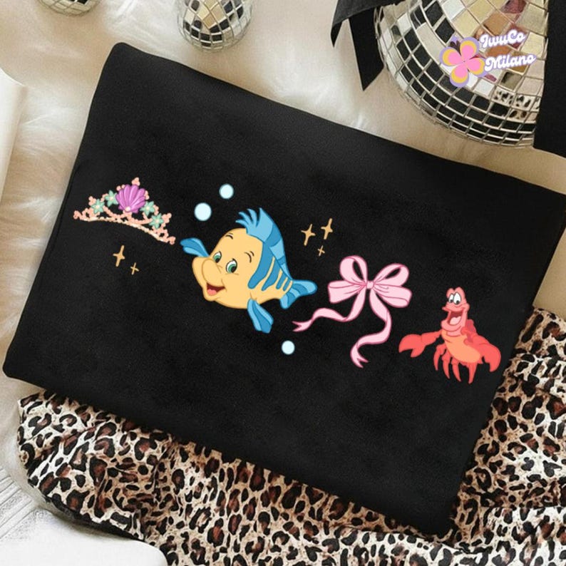 May include: Black blanket featuring colorful cartoon graphics of a tiara, Flounder, a pink bow, and Sebastian from The Little Mermaid. The blanket has a leopard print border.