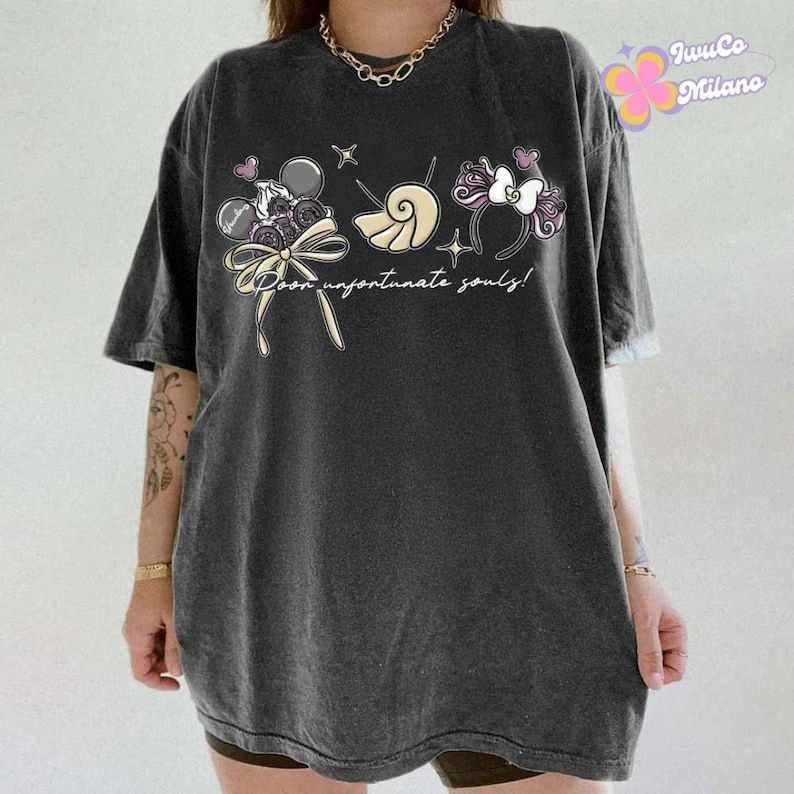May include: Dark gray t-shirt featuring a whimsical graphic with the text "Poor unfortunate souls!" The design includes cartoon elements like a bow, seashell, and a headband with a bow. The shirt has short sleeves and a relaxed fit.