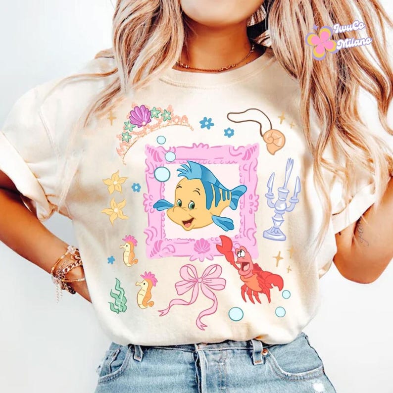 May include: Cream-colored t-shirt featuring a cartoon illustration of a yellow and blue fish inside a pink frame, surrounded by other sea-themed graphics. Additional elements include a crown, seahorses, and a red crab.