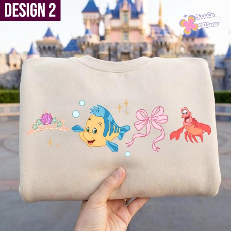 May include: Beige sweatshirt featuring a design with cartoon characters. The design includes a tiara, a yellow and blue fish, a pink bow, and a red crab. The text "DESIGN 2" is in the upper left corner.