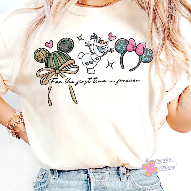 May include: A cream-coloured t-shirt with a playful design featuring Mickey Mouse ears, Olaf the snowman, and Minnie Mouse ears. The text "For the first time in forever" is written below the illustrations. The design includes hearts and stars.
