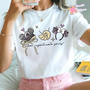 May include: White t-shirt with a whimsical graphic featuring a bouquet, seashell, and headband design. The text "Poor unfortunate souls!" is written in a script font. The design includes gray, pink, and cream colors.