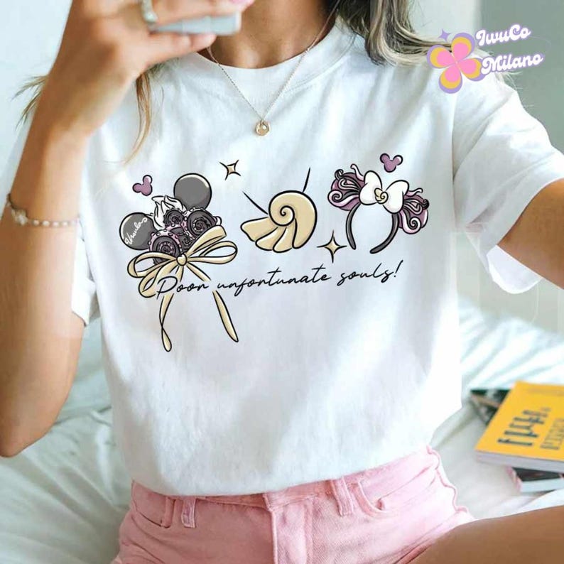 May include: White t-shirt with a whimsical graphic featuring a bouquet, seashell, and headband design. The text "Poor unfortunate souls!" is written in a script font. The design includes gray, pink, and cream colors.