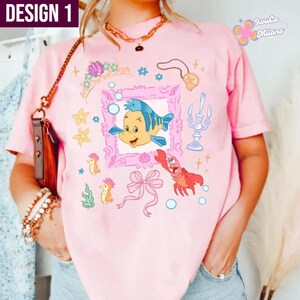 May include: Pink t-shirt featuring a cartoon illustration of a yellow and blue fish inside a pink frame, with other sea-themed graphics. The shirt has the text "DESIGN 1" in the upper left corner and the brand name "JuJuCo Milano" in the upper right corner.