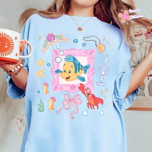 May include: Light blue t-shirt featuring a cartoon illustration of a yellow and blue fish inside a pink frame, surrounded by other sea-themed graphics. The shirt also has a pink bow, a red crab, and the text "JuwCo Milano".