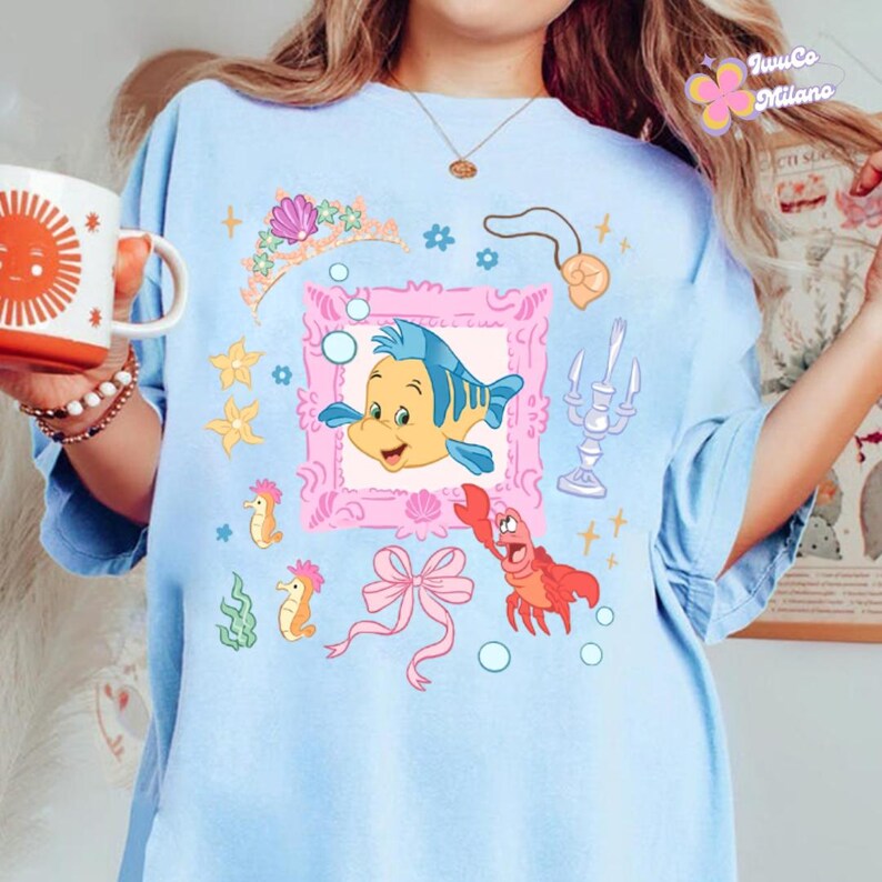 May include: Light blue t-shirt featuring a cartoon illustration of a yellow and blue fish inside a pink frame, surrounded by other sea-themed graphics. The shirt also has a pink bow, a red crab, and the text "JuwCo Milano".