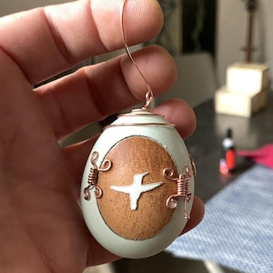 Może przedstawiać: Zielona, jajowata ozdoba z brązowym, drewnopodobnym wzorem i białą sylwetką kolibra. Ozdoba jest ozdobiona miedzianym drutem i ma miedzianą pętlę do zawieszenia.