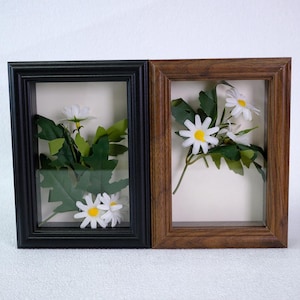 May include: Two framed floral arrangements. One frame is black, the other is brown. Each frame contains white daisies with yellow centers and green leaves. The frames are rectangular and have a shadow box effect.
