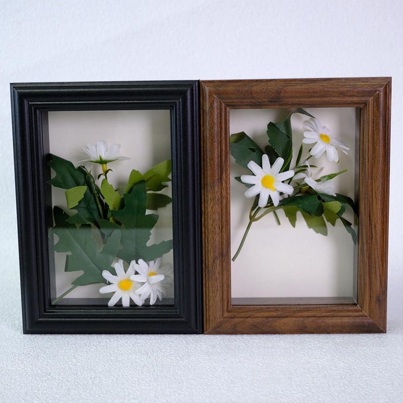 May include: Two framed floral arrangements. One frame is black, the other is brown. Each frame contains white daisies with yellow centers and green leaves. The frames are rectangular and have a shadow box effect.