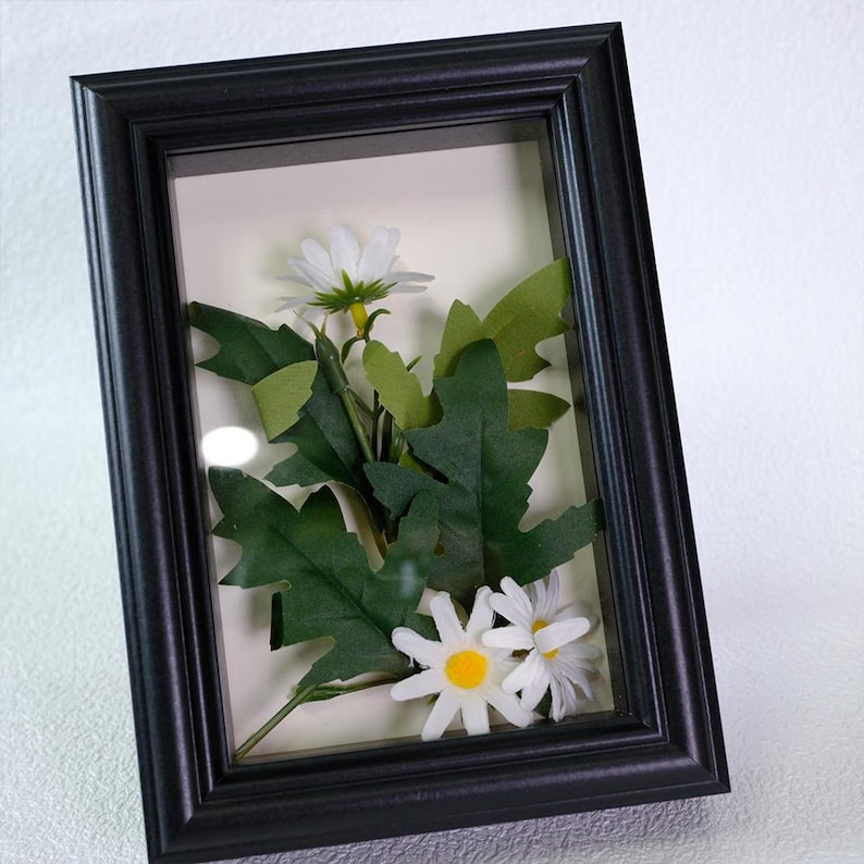 May include: A framed floral arrangement featuring white daisies with yellow centers and green leaves. The flowers and foliage are encased in a black rectangular frame with a glass front, creating a shadowbox effect. The background is a light cream color.