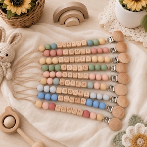 Puede incluir: Un conjunto de ocho sujetachupetes con cuentas de silicona de colores y cubos de madera con letras que forman nombres como Jamie, Bolle, Kaoly y Sarah. Cada uno tiene un broche de madera redondo con cierre metálico y un cordón.