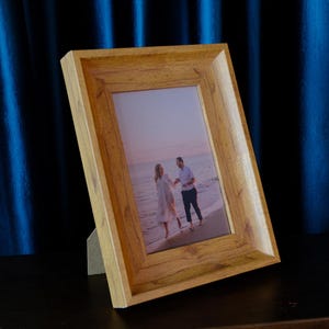 Op de afbeelding: Een rechthoekige houten fotolijst met een lichtbruine houtnerf afwerking. De lijst bevat een foto van een stel dat op het strand loopt. De lijst wordt ondersteund door een standaard.