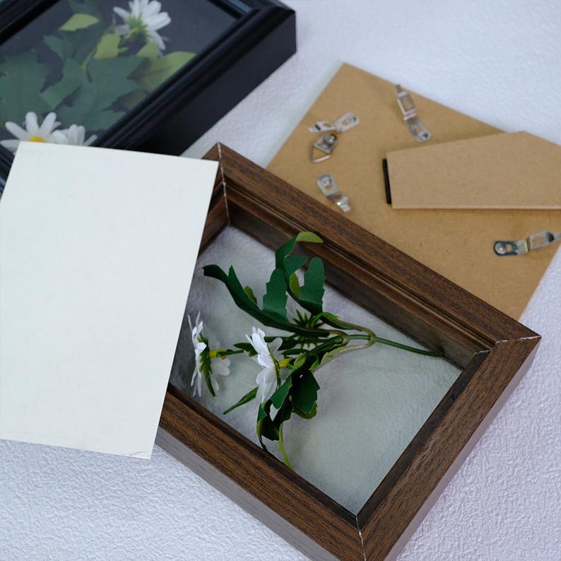 May include: A wooden shadow box frame with a sprig of white daisies and green leaves. A black shadow box frame with daisies is in the background. A blank white card and a cardboard backing with hanging hardware are also present.