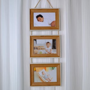 Peut inclure: Un présentoir photo à trois niveaux avec trois cadres en bois rectangulaires. Chaque cadre contient une photographie d'un bébé. Les cadres sont reliés par une corde naturelle et sont suspendus devant un rideau blanc.