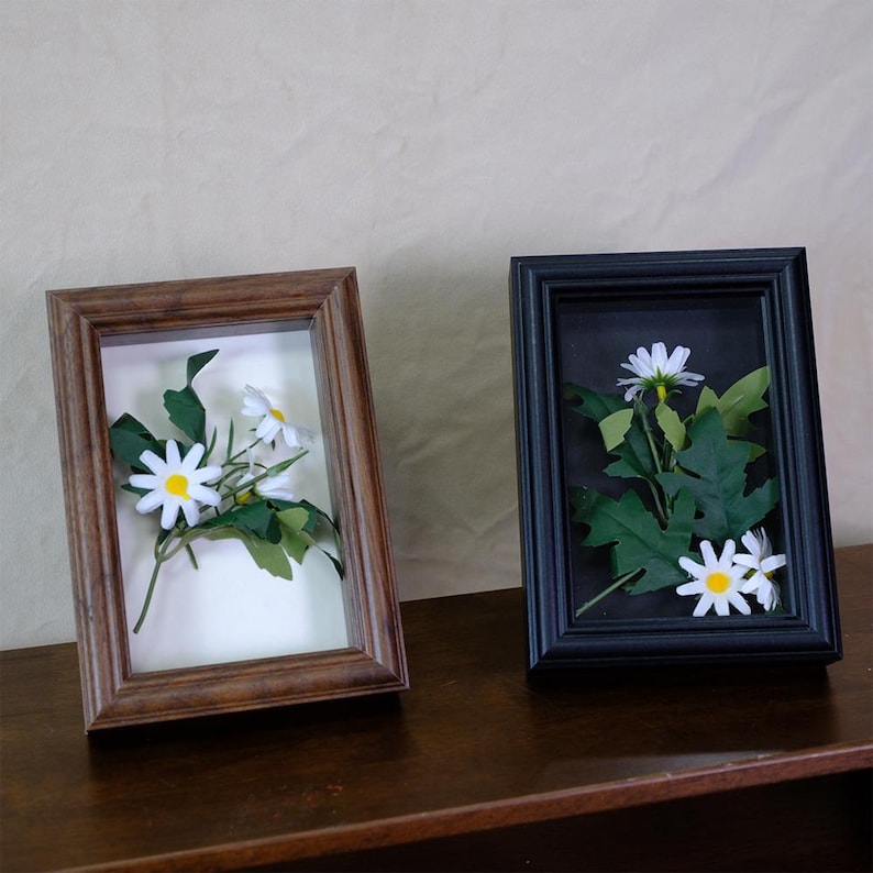 May include: Two framed floral arrangements. One frame is brown, the other is black. Each frame contains white daisies and green leaves. The frames are rectangular and sit on a dark brown surface.
