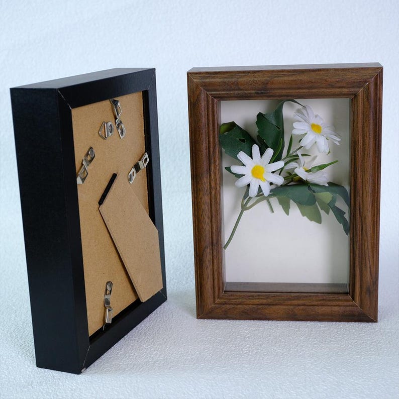 May include: Two rectangular picture frames. One is black, with the back and stand visible. The other is a brown wooden frame, displaying white daisies with yellow centers and green leaves against a white background. The frames are side by side.