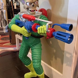 May include: A green and yellow alien figure with a large head and a brain-like texture. The alien is holding a blue and red toy gun with a long barrel. The figure is standing on a wooden floor.