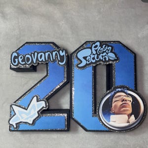 May include: A blue and black number 20 birthday decoration with silver glitter trim. The number 2 has the name "Geovanny" in white script. The number 0 has the words "Playa Saturn" and a photo of a person.