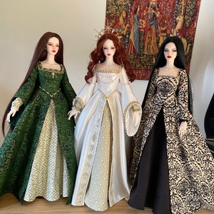 May include: Three fashion dolls wearing elaborate gowns. The doll on the left is wearing a green gown with gold accents. The doll in the middle is wearing a white gown with gold accents. The doll on the right is wearing a black gown with gold accents.