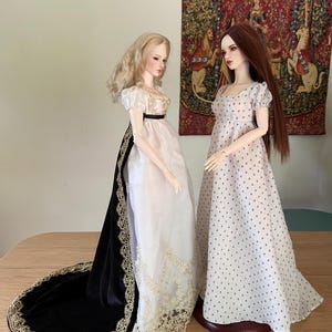 May include: Two dolls in period gowns. One doll wears a white dress with a black sash and a long black velvet train with gold trim. The other doll is in a white dress with a small black pattern. Both dolls have long hair and are standing on a wooden table.