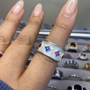 May include: A silver ring with a white enamel band and red and blue square gemstones. The ring is on a finger with clear nail polish.