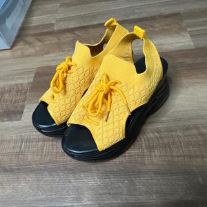 May include: A pair of bright yellow knit platform sandals featuring an open-toe design, a lace-up front with matching yellow laces, and a thick black rubber sole. The upper portion is constructed from a textured, diamond-patterned fabric.