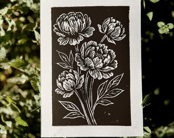 May include: A black and white linocut print featuring a bouquet of peonies. The floral design is rendered in white against a black background, with detailed petals and leaves. The print is set within a white border.