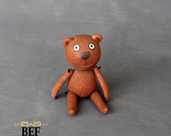 Puede incluir: Un oso de peluche marrón con ojos verdes y una sonrisa pintada se sienta sobre una superficie gris. El oso tiene una superficie texturizada y está hecho de madera.