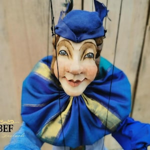 Op de afbeelding: Een blauwe en gele pop met een geschilderd gezicht en een blauwe hoed. De pop is een nar met een brede glimlach en grote ogen.