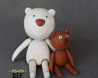 Puede incluir: Dos osos de peluche de madera hechos a mano, uno blanco con ojos azules y nariz marrón, y otro marrón con ojos verdes. Los osos están sentados juntos, con el oso blanco a la izquierda y el oso marrón a la derecha.