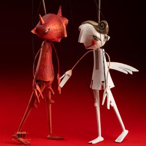 May include: Two wooden marionette puppets, one red devil with horns and one white angel with wings, stand against a red background. The devil has a pointed tail and the angel is reaching out to the devil.