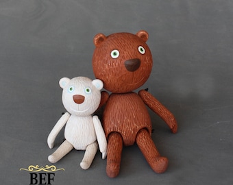 Puede incluir: Dos osos de peluche de madera hechos a mano. El oso más grande es marrón con ojos verdes y una nariz marrón. El oso más pequeño es blanco con ojos azules y una nariz marrón. Ambos osos están sentados con los brazos extendidos.