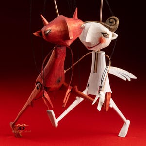 May include: Two wooden marionette puppets. One is red with horns, the other is white with a swirl of hair. Both have jointed limbs and are suspended by strings against a red background. The red puppet has a devilish appearance, while the white puppet has a more angelic look.