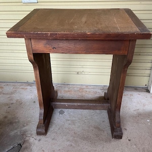 May include: A brown wooden desk with a rectangular top and a slanted surface. The desk features two vertical side supports connected by a horizontal base stretcher. The wood shows visible grain patterns and signs of wear on the surface edges.