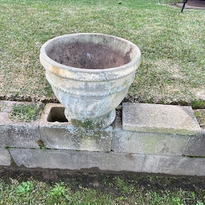 May include: A weathered, light grey stone planter with a wide, circular opening and a thick, rounded rim. The exterior surface features a textured, aged finish with visible signs of moss and discolouration, resting on a pedestal base.