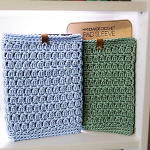 Pode incluir: Duas capas para iPad de crochê feitas à mão, uma azul claro e outra verde, em uma prateleira branca com livros atrás. A placa acima das capas diz "Handmade Crochet iPad Sleeve".
