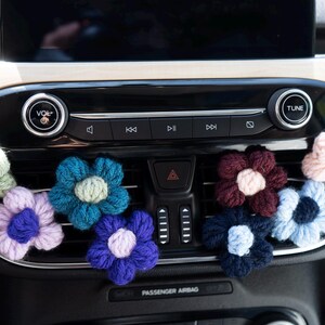 Könnte beinhalten: Eine Sammlung bunter, gehäkelter Blumen-Autolüftungsclips. Die Blumen sind in verschiedenen Blau-, Lila-, Rosa- und Grüntönen gehalten, mit kontrastierenden Mittelfarben. Sie sind an einer schwarzen Autolüftung befestigt.