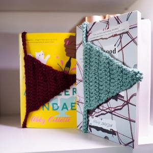 以下が含まれることがあります： 手作りのかぎ針編みのブックマークが付いた2冊の本。1冊はマルーンのブックマークが付いた黄色、もう1冊はティールのブックマークが付いた白です。本は白い棚にあります。本は読書用です。