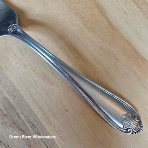 May include: Close-up of a silver fork with a decorative floral design on the handle and tines. The fork is made of a shiny, reflective metal and is set against a light-coloured wooden surface. The fork is a piece of silverware.