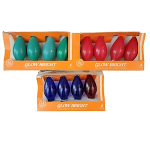May include: Three boxes of GE Glow Bright outdoor light bulbs. Each box contains four bulbs in different colours: teal, red, blue, and burgundy. The boxes are orange and white with the brand name and bulb count printed on them.