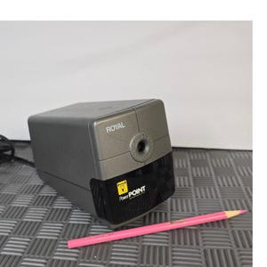 Puede incluir: Un sacapuntas eléctrico gris con la palabra "ROYAL" impresa en la parte superior. El sacapuntas tiene una base negra con el logotipo "PowerPOINT". Un lápiz rosa está sobre la superficie.